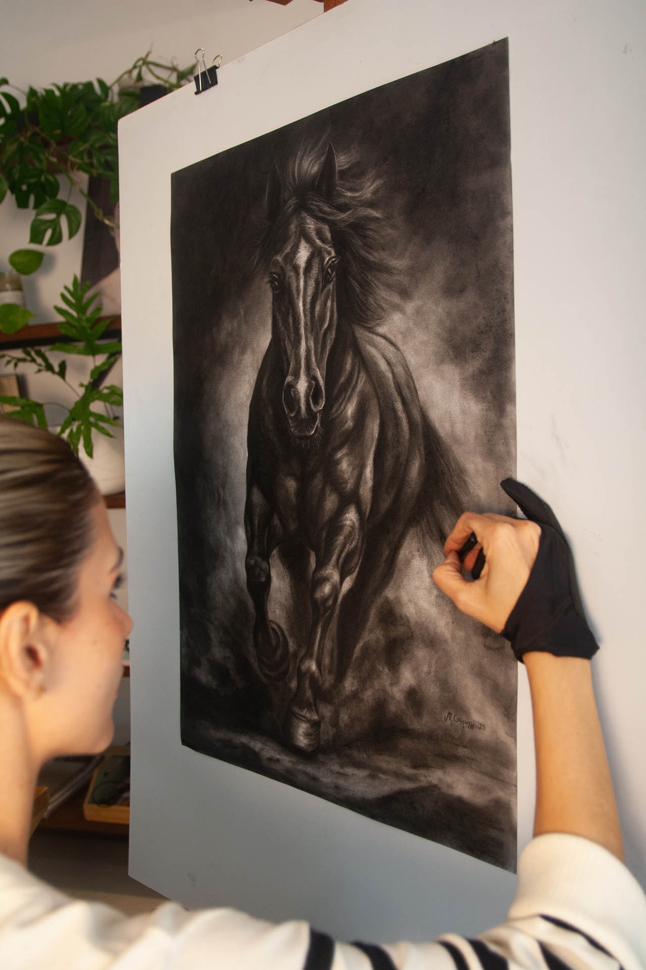 Mariela Crapuzzi de espaldas, trabajando con la mano enguantada sobre un carboncillo de un caballo al galope montado en la pared del estudio.