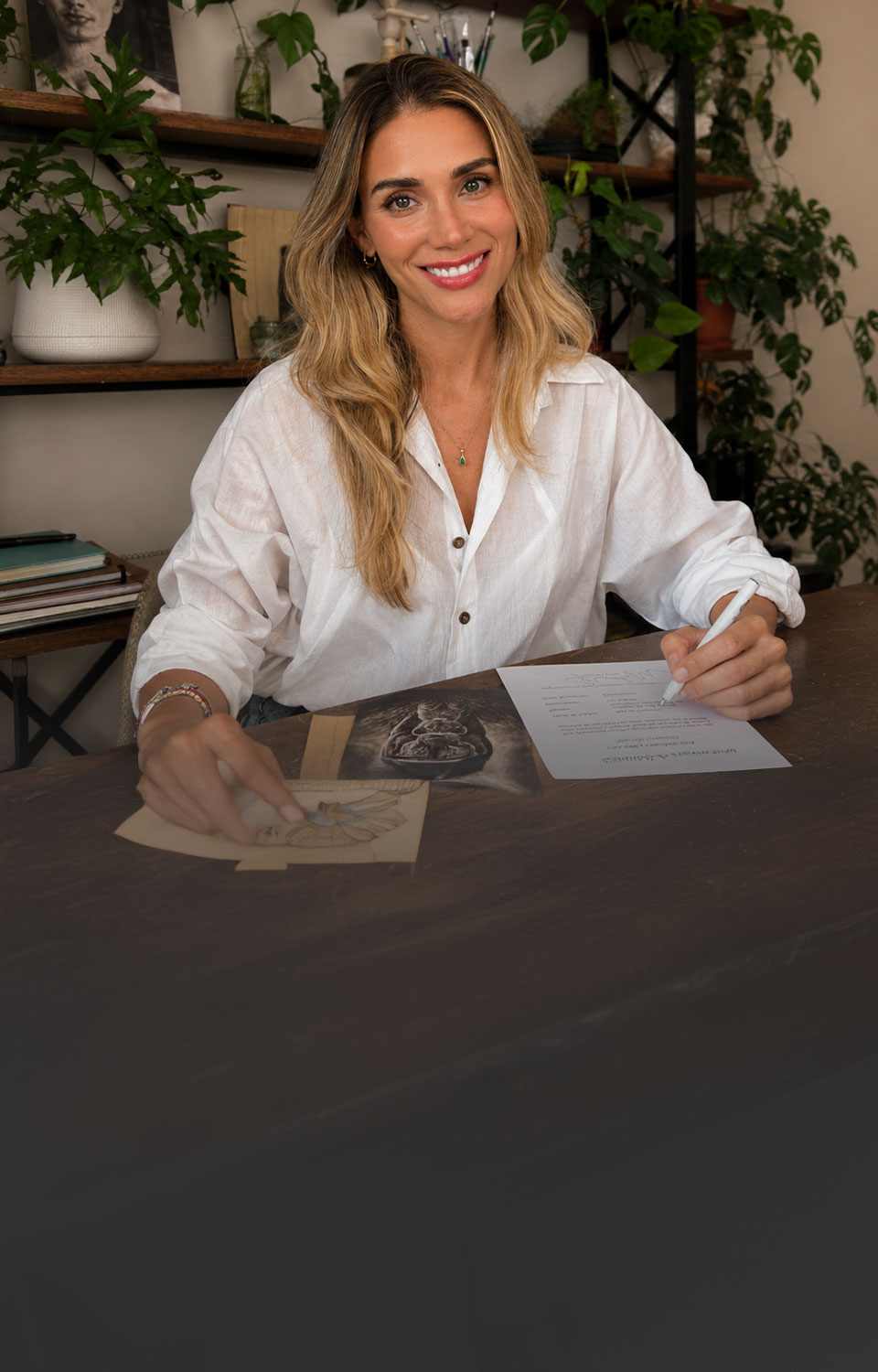 Mariela Crapuzzi sentada a su mesa de trabajo, firmando el certificado de autenticidad de una obra, con estanterías de plantas y materiales de dibujo detrás.